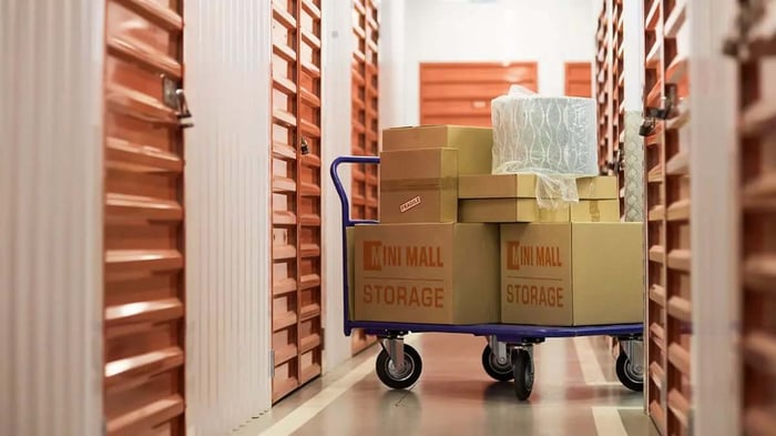 a storage facility hallway filled with climate controlled storage units and a hand cart covered in Mini Mall Storage branded moving boxes
